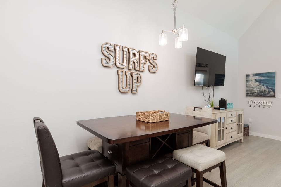Your stylish dining space features coastal charm with 'SURF'S UP' wall art and a warm wood table perfect for family meals.
