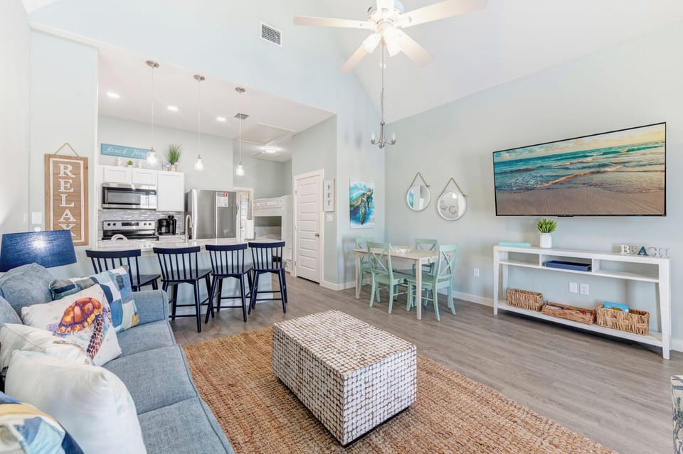 Relax in your bright, coastal-themed living space with ocean artwork, comfy seating, and modern kitchen—perfect for beach getaway memories.