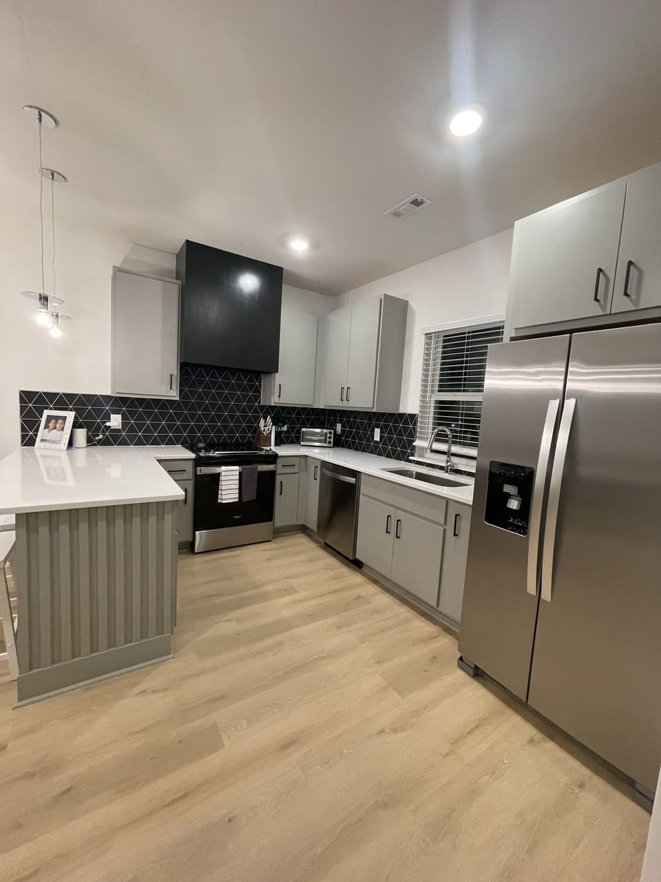  KITCHEN – This sleek, fully equipped kitchen features stainless steel appliances, elegant gray cabinetry, and a striking black geometric backsplash. The island with bar seating offers a convenient space for quick meals or entertaining. Pendant lights and recessed lighting brighten the room, highlighting the clean lines and contemporary design. Whether you're preparing a gourme...