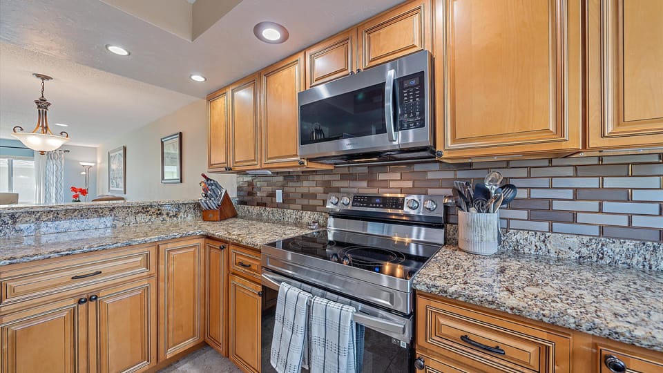 A modern kitchen with wooden cabinets, granite countertops, stainless steel microwave and oven, black and gray tiled backsplash, and utensils in a holder near the stove.