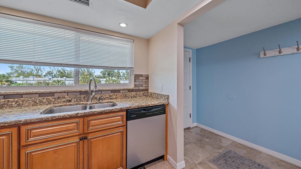 A kitchen interior featuring a stainless steel sink and dishwasher set into a granite countertop with light brown cabinetry. The right wall is light blue with a few hooks for hanging items.
