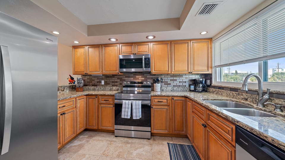 Modern kitchen with wooden cabinets, stainless steel appliances, granite countertops, double sink, and a tile backsplash. There is a rug on the tiled floor and a window with blinds over the sink.
