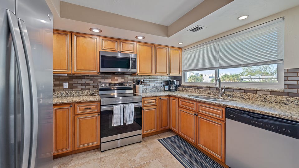 A modern kitchen with wooden cabinets, stainless steel appliances, granite countertops, tiled backsplash, and a window with blinds. A rug is placed in front of the sink and dishwasher.