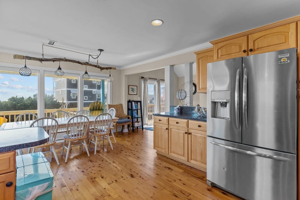 Oak Island, NC vacation rental kitchen featuring stainless French-door refrigerator with ice maker natural wood cabinets, dark countertops, hardwood floors, coastal decor, and an open view into the living area with sliding glass doors and deck beyond