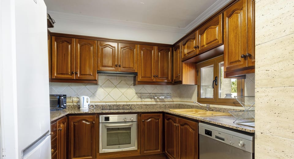Fully equipped kitchen at Villa Alejandra featuring wooden cabinets, oven, dishwasher, fridge, spacious worktop and a window providing natural light.