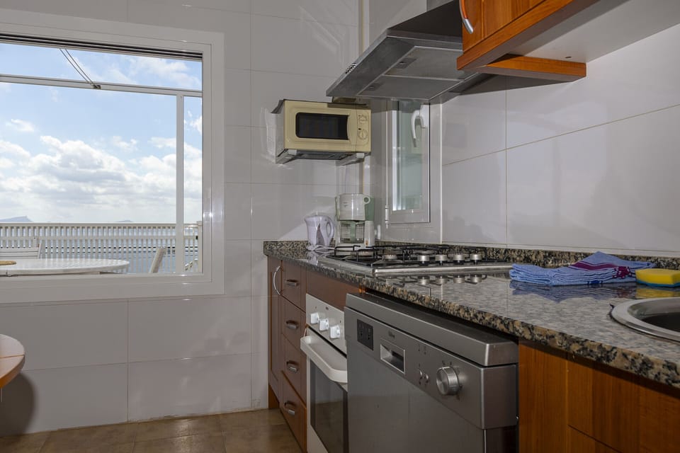 Side view of Villa Maria’s fully equipped kitchen, featuring oven, dishwasher, microwave and spacious countertops. A practical and bright space with a window and visual access to the outdoor terrace.
