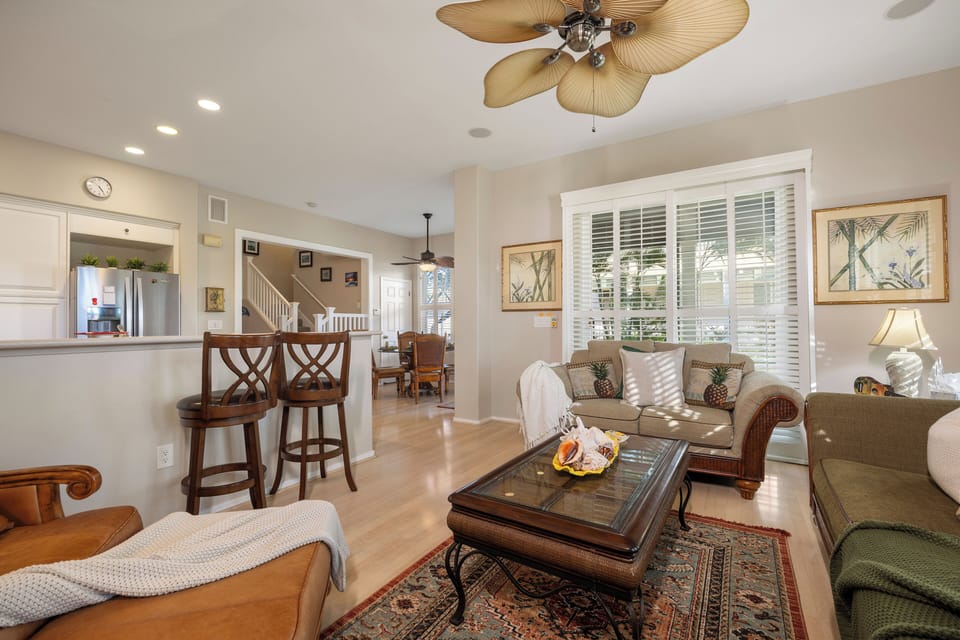 Relax in your tropical-themed living space featuring comfortable seating, tropical artwork, and an open layout connecting to the kitchen.