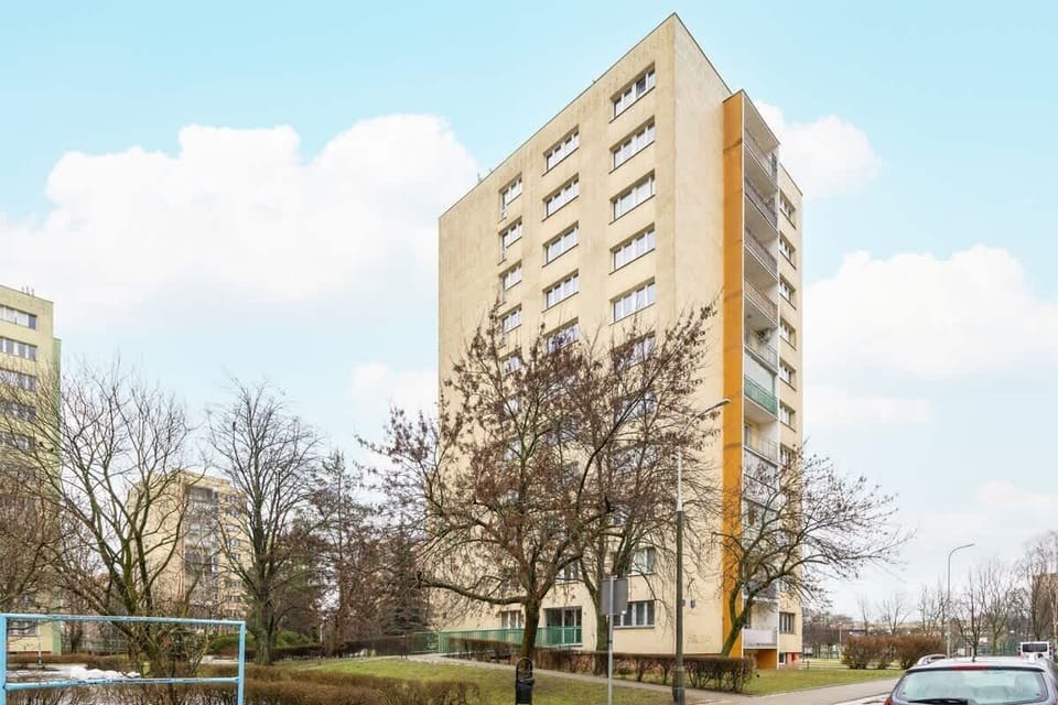 A towering residential apartment building with a bright yellow facade, surrounded by trees and an open area, offering a lively urban living experience.







