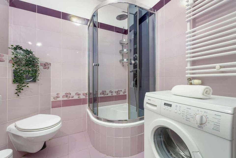 A spacious bathroom with pastel pink tiles, a sleek glass shower cabin, a modern washing machine, and a compact yet practical vanity unit.
