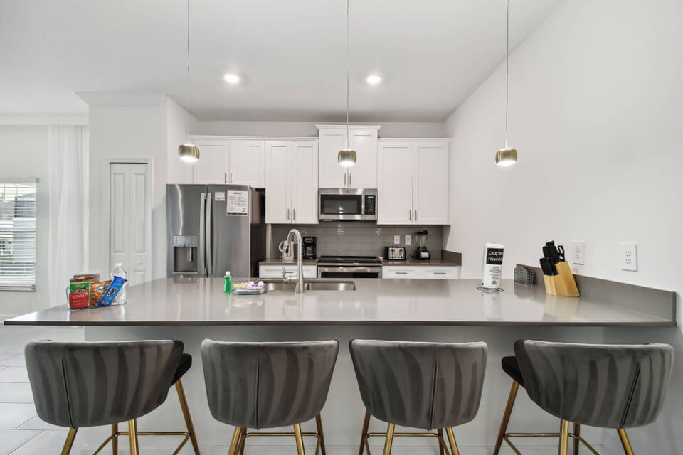 Enjoy a stylish kitchen featuring a spacious gray countertop and cozy bar stools for casual meals. Bright lighting enhances modern stainless steel appliances and sleek white cabinetry, perfect for all your culinary creations.