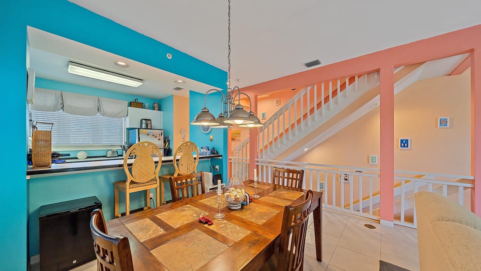 A brightly colored dining area with a wooden table and six chairs next to an open kitchen. The space features vibrant blue and coral walls, white stairs, and modern pendant lighting.