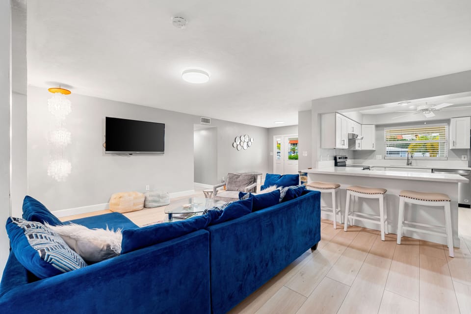 Modern living room with a blue sofa, wall-mounted TV, and a minimalistic coffee table. The room transitions into an open-plan kitchen with white cabinetry and a breakfast bar with stools.