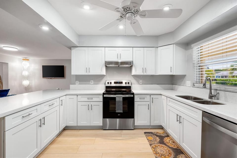 A modern kitchen with white cabinets, stainless steel appliances, a black stove, double sink, and a vent hood. There is a ceiling fan, a wall-mounted TV, and a window with blinds over the sink.