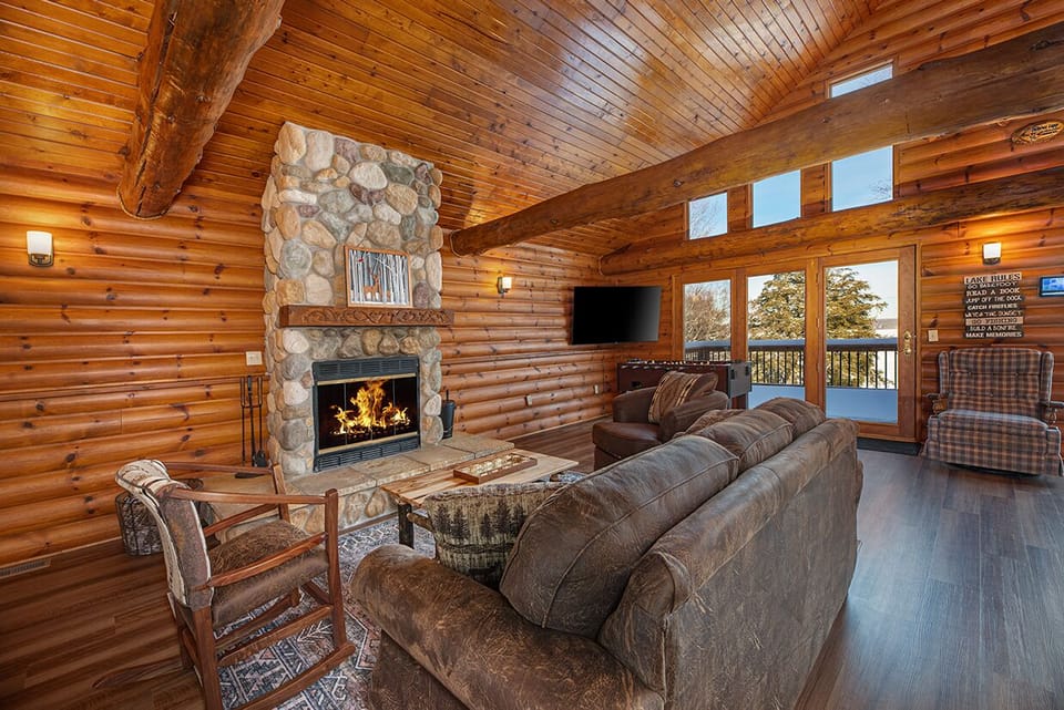 - Warm log interior with stone fireplace and comfy seating - Forest views through tall windows brighten the space - Rustic living room ideal for gatherings and calm nights