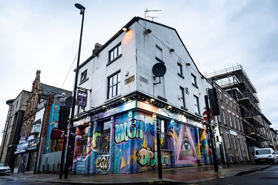 This striking street-art scene captures the creative spirit surrounding Camden Apartments. A bold mural featuring a vivid blue eye set within a pink triangle takes centre stage, framed by bursts of colour and contemporary graffiti lettering. Reflected perfectly in a rain-kissed pavement below, the image creates a dramatic mirror effect that feels effortlessly cool and unmistakably city-centric.