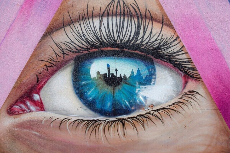 This striking street-art scene captures the creative spirit surrounding Camden Apartments. A bold mural featuring a vivid blue eye set within a pink triangle takes centre stage, framed by bursts of colour and contemporary graffiti lettering. Reflected perfectly in a rain-kissed pavement below, the image creates a dramatic mirror effect that feels effortlessly cool and unmistakably city-centric.