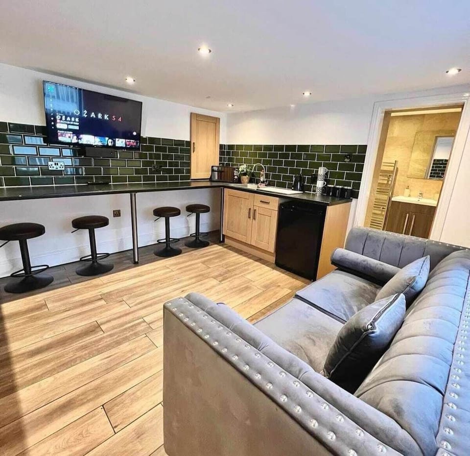 A modern, open-plan living space designed for comfort and convenience.

The stylish kitchenette features sleek dark green tiled splashbacks, wooden cabinetry, and a practical worktop area fully equipped with air fryer, toaster, microwave, kettle and fridge — perfect for everything from quick breakfasts to relaxed evening meals.

A wall-mounted Smart TV sits above the breakfast bar, so you can stream your favourite shows while cooking or unwinding. The breakfast bar with comfortable stools creates an ideal space for casual dining, morning coffee, or catching up after a busy day.

The room flows into a cosy seating area with a plush sofa, offering a comfortable spot to relax, watch TV, or socialise. Bright, clean, and thoughtfully laid out, this space blends practicality with modern comfort for a stress-free stay.