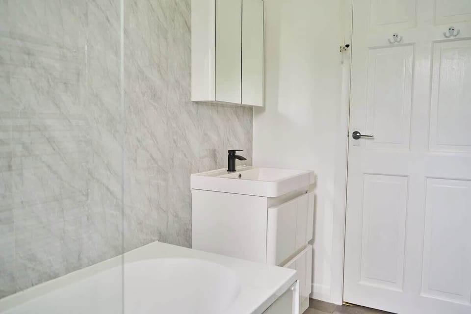 Step into this clean bathroom setup featuring white marble-effect wall paneling, a compact white vanity with a black faucet, a mirror cabinet, and a bathtub.
