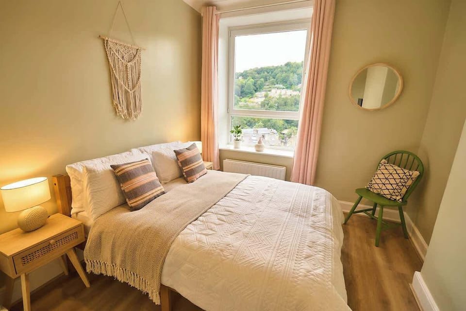 Tour this inviting bedroom featuring soft pink curtains framing a view of lush green hills, macramé wall hanging, a green accent chair, and warm bedside lighting.