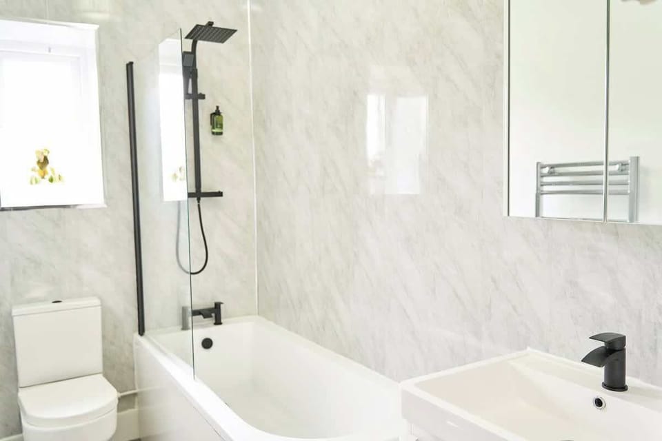 View the full bathroom layout showcasing a white bathtub with a matte black shower fixture, marble-effect wall paneling, a toilet, and a vanity sink with a mirror cabinet.