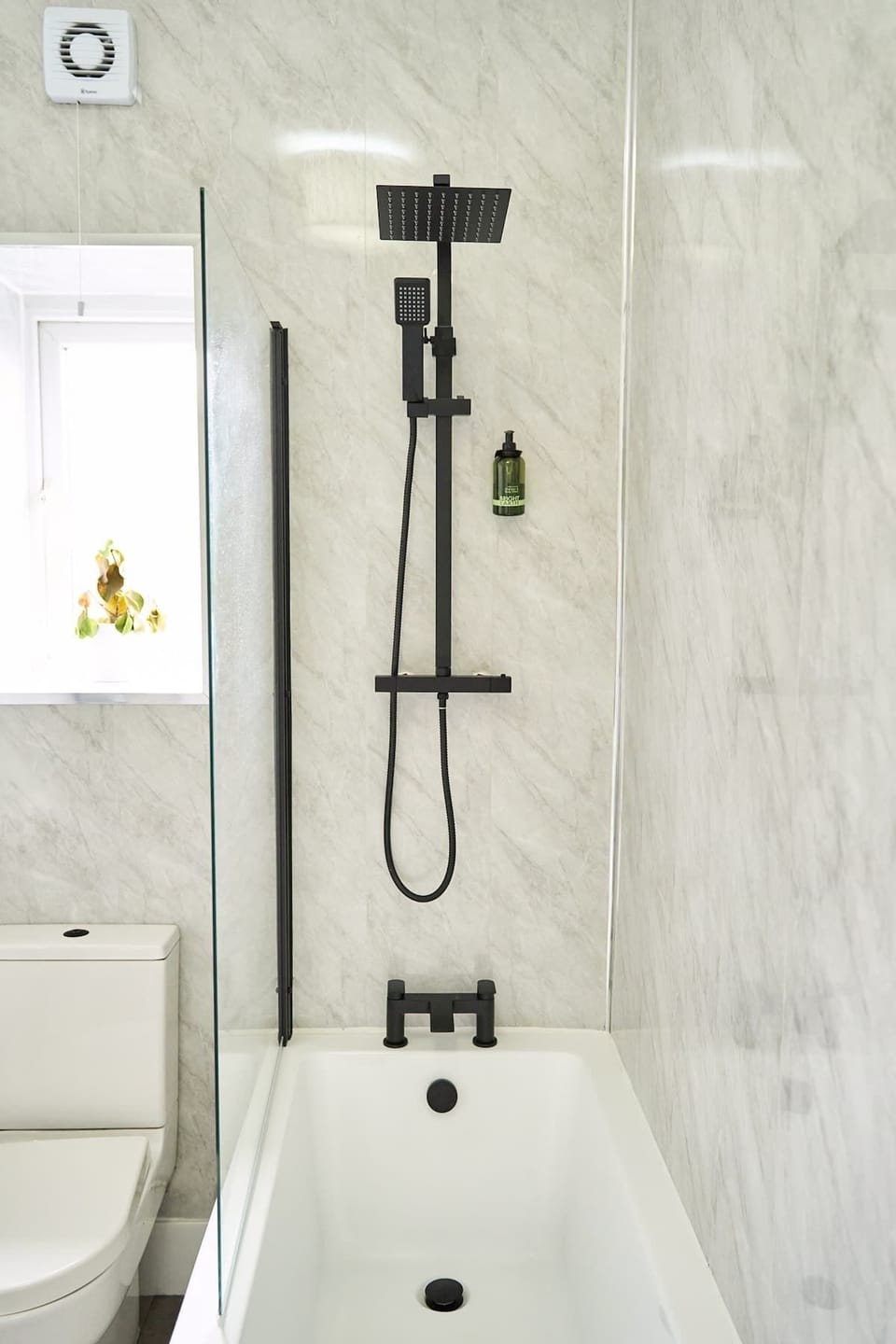Detail the shower-over-bath setup styled with matte black fixture hardware, including a rainfall showerhead, a handheld sprayer, and a glass screen panel.