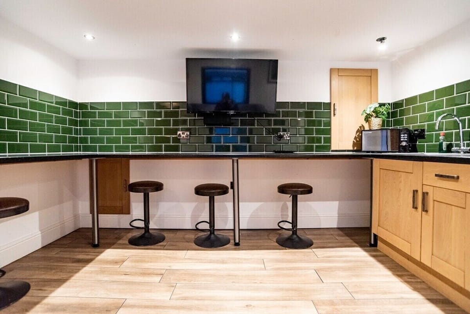 and sofa and breakfast bar chairs

A modern, open-plan living space designed for comfort and convenience.

The stylish kitchenette features sleek dark green tiled splashbacks, wooden cabinetry, and a practical worktop area fully equipped with air fryer, toaster, microwave, kettle and fridge — perfect for everything from quick breakfasts to relaxed evening meals.

A wall-mounted Smart TV sits above the breakfast bar, so you can stream your favourite shows while cooking or unwinding. The breakfast bar with comfortable stools creates an ideal space for casual dining, morning coffee, or catching up after a busy day.

The room flows into a cosy seating area with a plush sofa, offering a comfortable spot to relax, watch TV, or socialise. Bright, clean, and thoughtfully laid out, this space blends practicality with modern comfort for a stress-free stay.