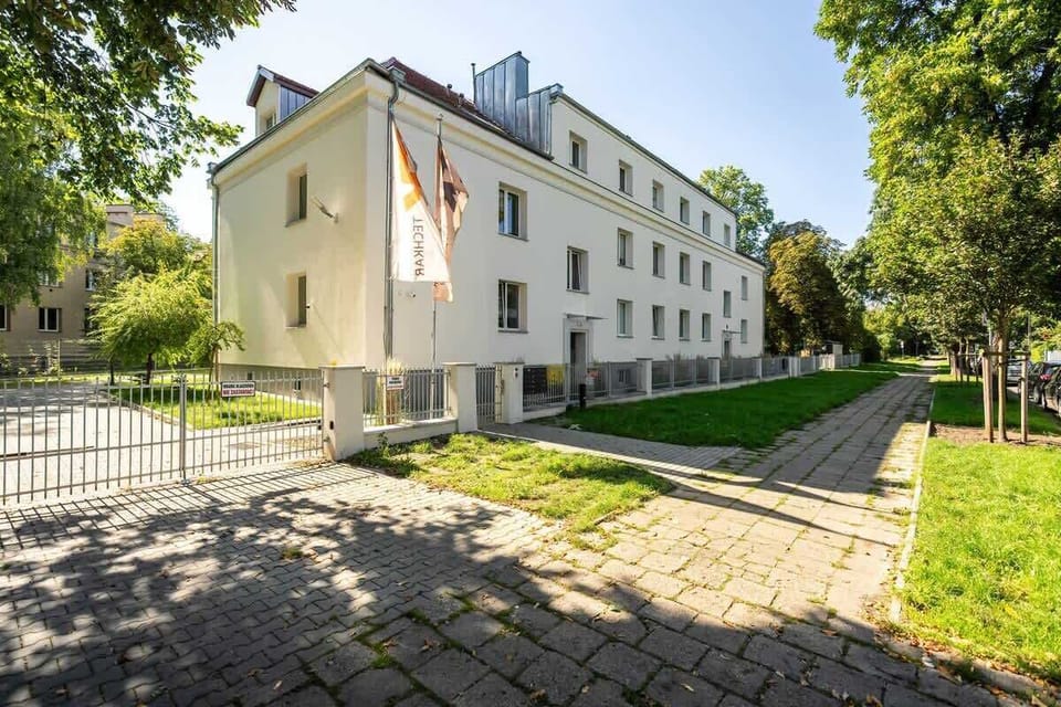 The exterior of a residential building with a green courtyard, trees, and a paved walkway.
