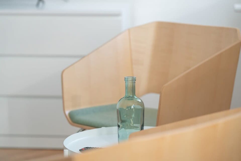 A close-up of a wooden chair with a green cushion, placed near a bedside table.
