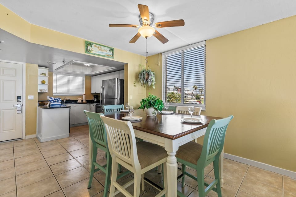 This charming dining space offers a comfortable setting with ample sunlight through large windows. The hanging plant and light fixtures add a relaxed, tropical vibe, while the open layout leads to a fully-equipped modern kitchen.