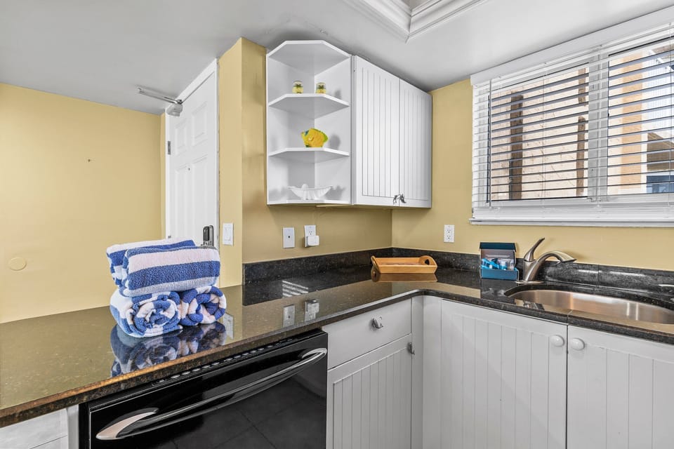 This kitchen area features a sleek granite countertop, open shelving for easy access, and modern cabinetry. Its efficient design is perfect for meal preparation and includes a deep sink with natural light pouring in from nearby windows.