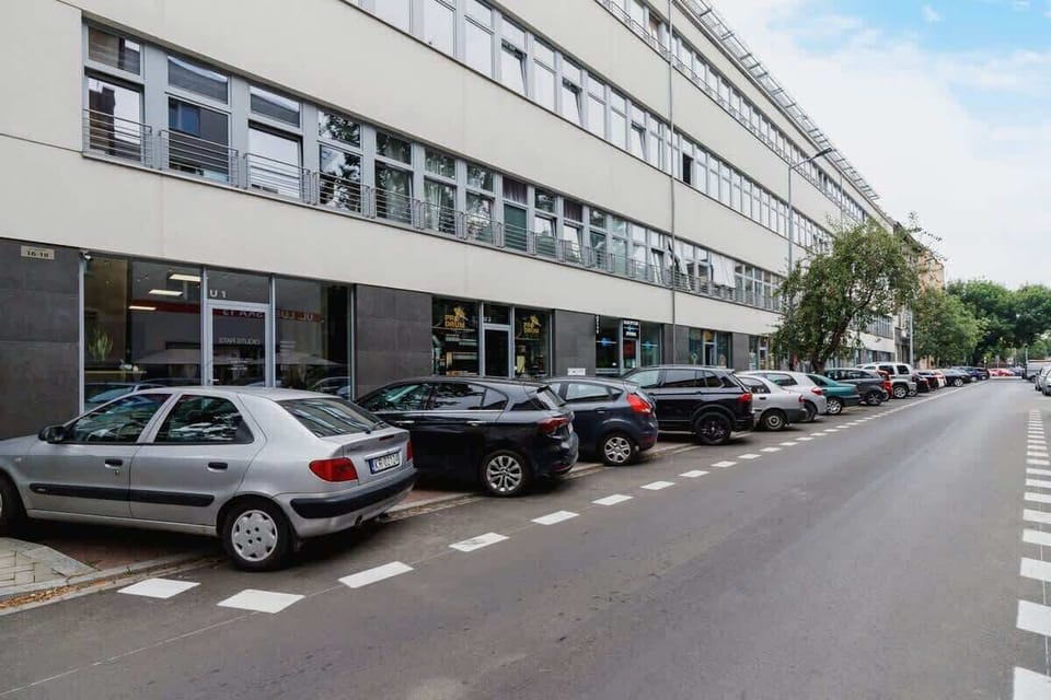 A lively street view with parked cars, a clean sidewalk, and a modern white building with large windows, contributing to an urban and accessible setting.






