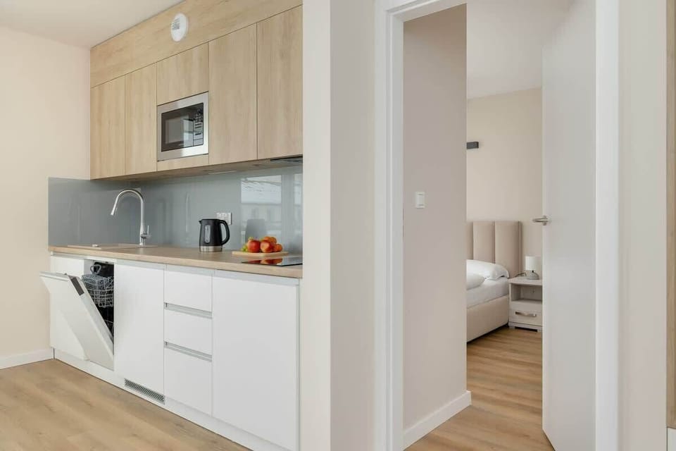 A compact yet functional kitchen with light wooden cabinets, a gray backsplash, and an integrated oven. A glimpse of the hallway leading to other rooms.

