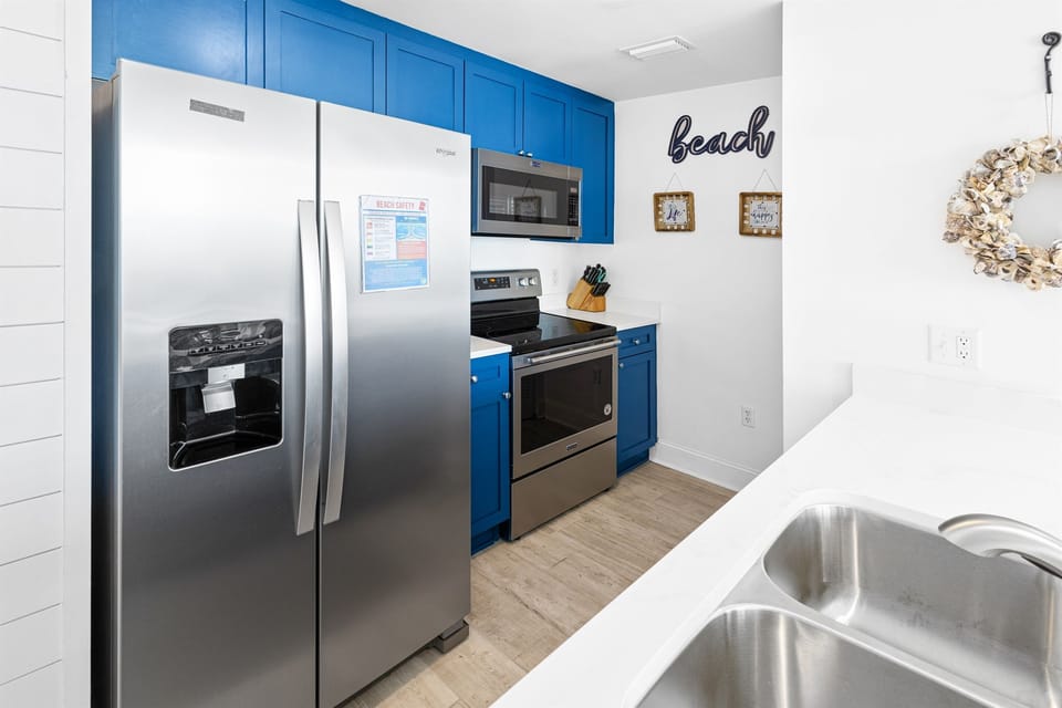 Your bright, beach-themed kitchen features vibrant blue cabinets and stainless steel appliances, creating a cheerful coastal atmosphere for your vacation cooking.