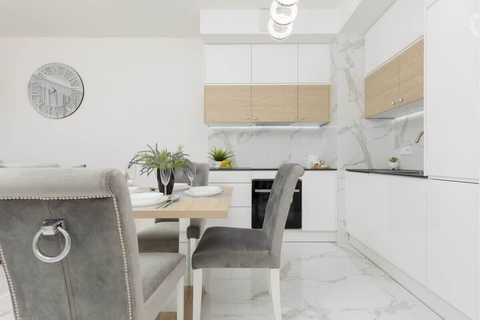 A dining area connected to the kitchen, with a glass table, grey velvet chairs, and decorative plants, maintaining a classy and contemporary style.
