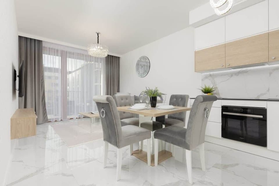 An open-plan kitchen and dining area with light wood cabinets, modern appliances, and a glass dining table with grey chairs.
