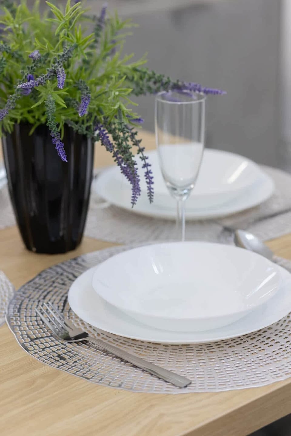 A close-up of a dining table decorated with elegant tableware, a glass vase with green and purple plants, and woven placemats.
