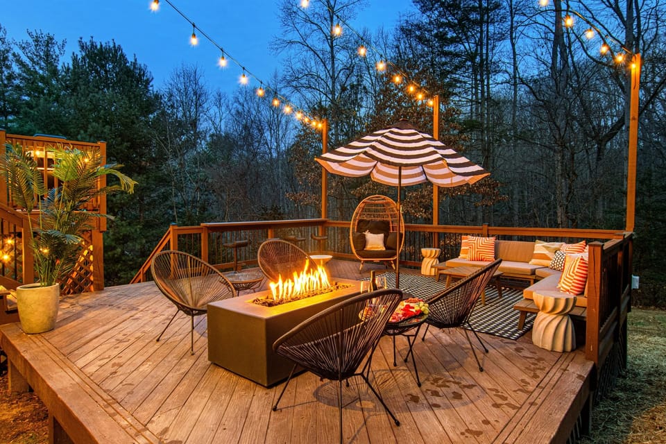 The lower deck at dusk — gas fire pit, string lights, lounge chairs, plush sofa, and an egg chair. This is where your group ends up every evening.
