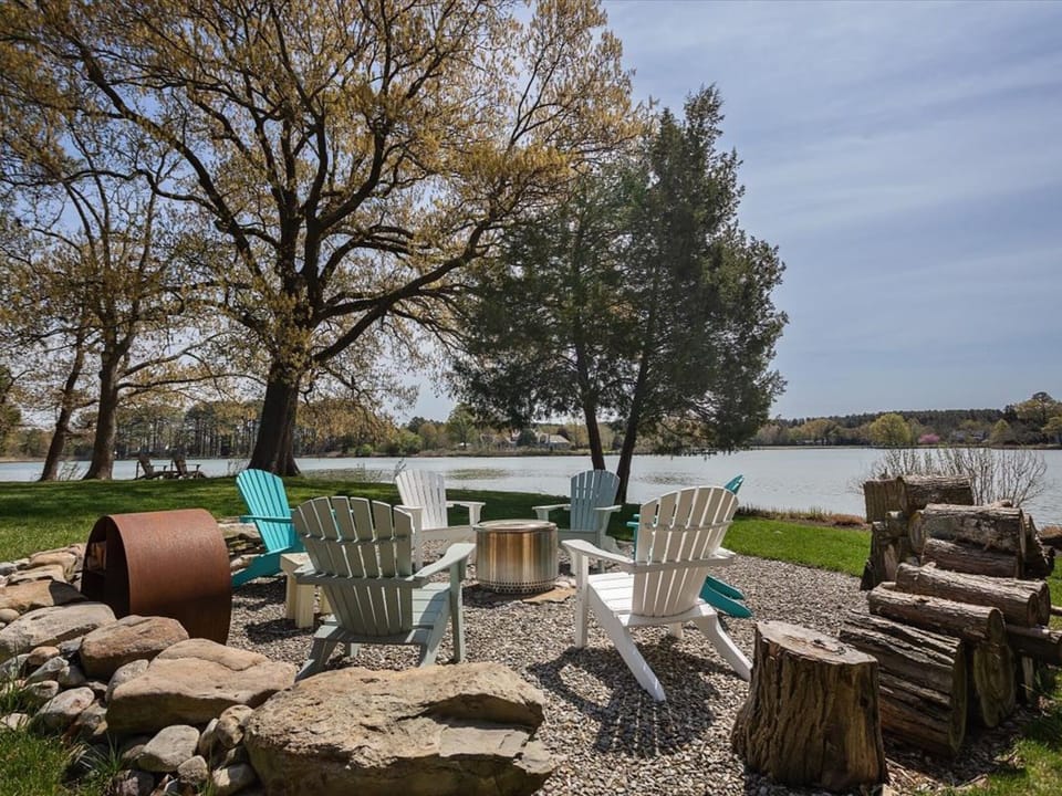 Peaceful lakefront seating area with Adirondack chairs, fire pit, and stacked firewood overlooking calm waters surrounded by seasonal trees.