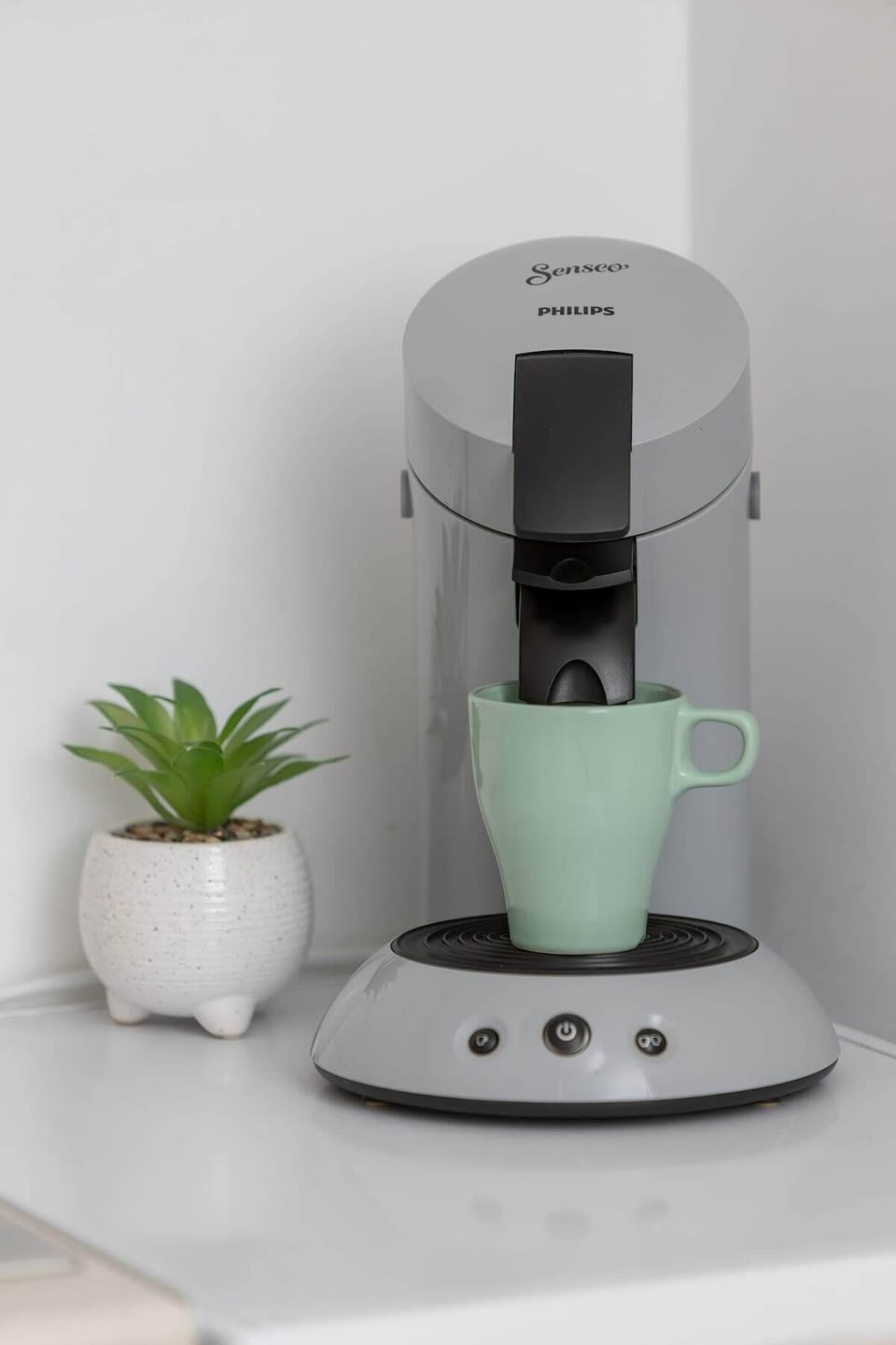 A close-up of a modern coffee machine, a small green plant, and a neutral wall, adding a sleek touch to the kitchen.

