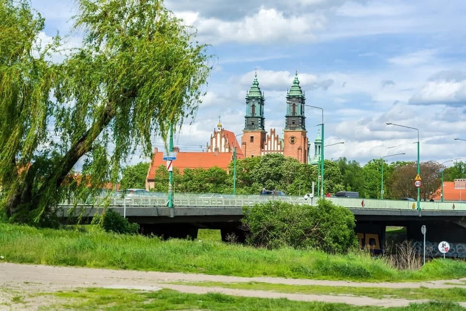 A scenic riverside view with lush greenery, a walking path, and a bridge leading towards a beautiful historic church with twin towers in the background.
