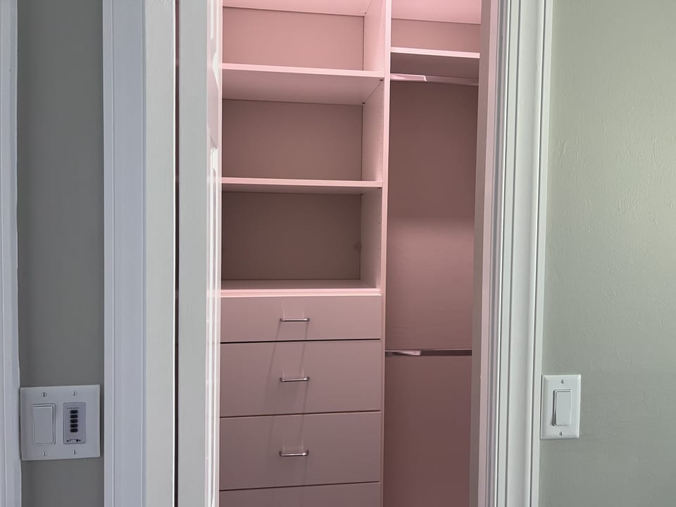 Small, empty walk-in closet with pink built-in shelving and drawers, seen through an open door. Light switches are visible on the adjacent wall.