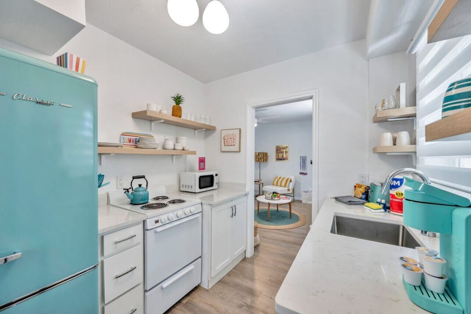 Bright kitchen view w/ open shelving, teal accents, & a clean, airy layout.