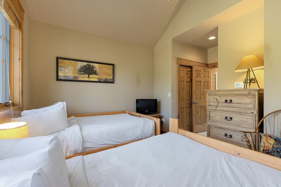 A bedroom with two twin beds, a dresser with a lamp, a small TV on a stand, and a framed painting of a tree on the wall. The room has wooden furniture and a door leading to another area.