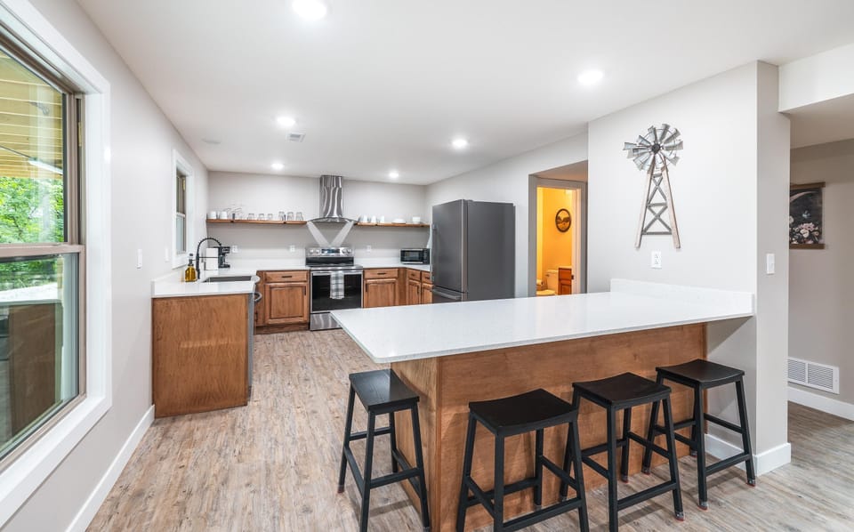 4 person seating at island and kitchen view.  Area has lots of lower cabinet space, open shelving above countertops.  All stainless steel appliances including stove, refrigerator, dishwasher, microwave, and large stainless steel farm sink.