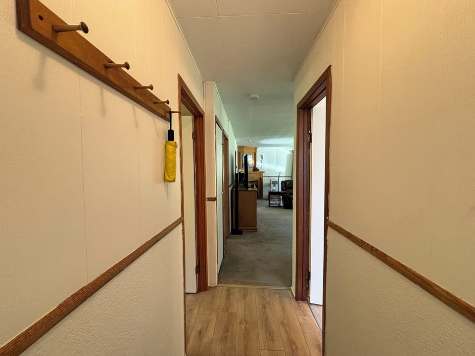 A hallway with neutral walls and wood trim, leading to a living area. A wooden coat rack. The hallway provides access to multiple rooms and opens up into a spacious living area creating a welcoming pathway through the home.