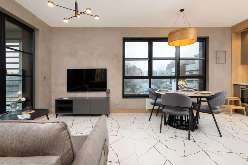 A spacious living area with a gray sofa, a TV, a dining table, and large windows offering a panoramic city view.

