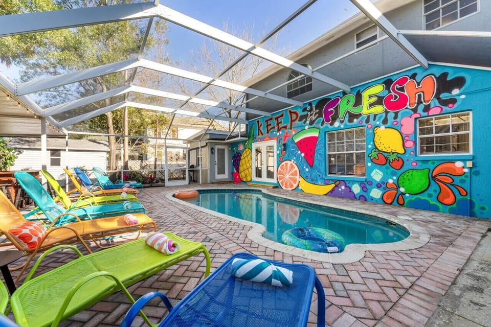 Featuring a stunning outdoor scene, with a large swimming pool as the focal point. The pool is surrounded by colorful chairs and sun loungers, creating an inviting and relaxing atmosphere. The vibrant mural painted on its side adds an artistic touch to th