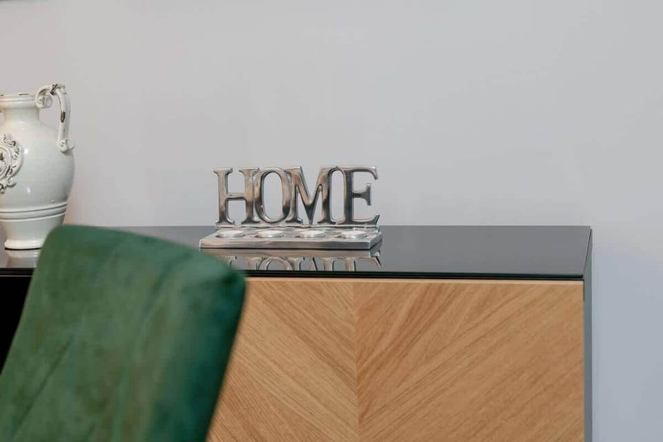 A close-up of a wooden sideboard with a decorative "HOME" sign and a plush green chair partially visible in the frame.

