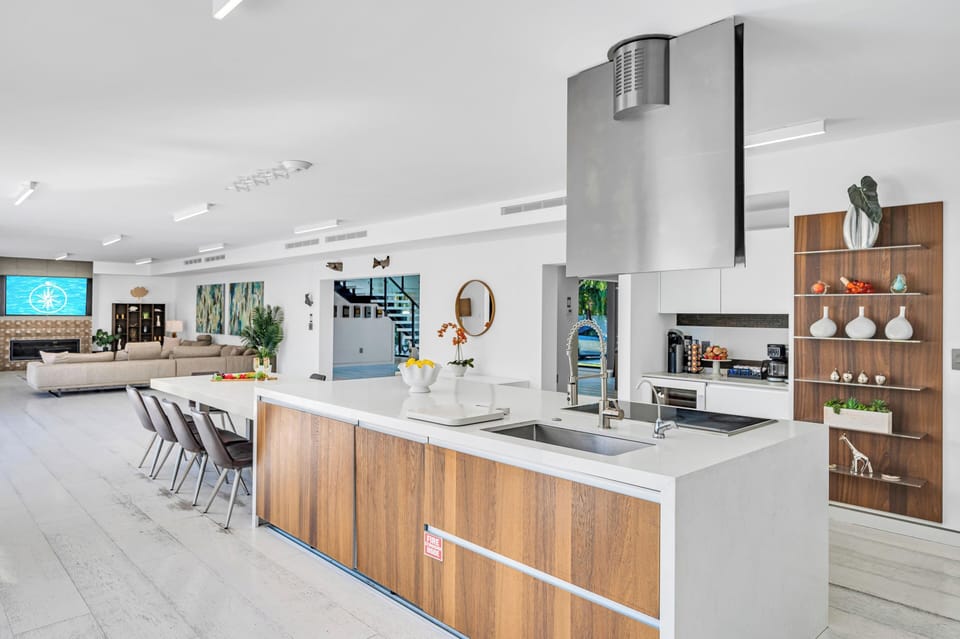 The kitchen is lined with sliding floor-to-ceiling windows, offering waterfront views and seamless access to the pool and outdoor kitchens.