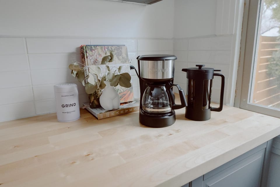 A kitchen counter with a coffee maker, a French press, a small plant, a candle labeled "GRIND," and a few books near a window.